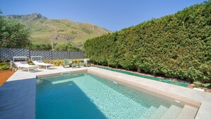 Une piscine rectangulaire à l’eau d’un bleu limpide, entourée d’une haie basse, et un espace détente avec des chaises longues et une table.