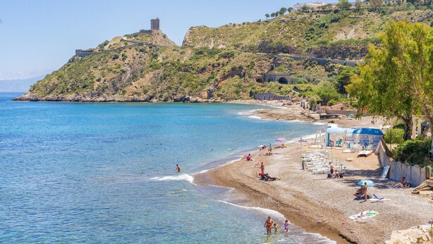 Une plage à l’eau d’un bleu limpide, une rive sablonneuse et, au loin, une colline surmontée d’une tour.