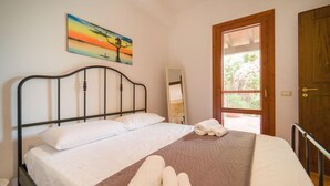 Un lit avec du linge de lit blanc, une couverture marron et des serviettes enroulées. Un tableau représentant un arbre et un coucher de soleil est accroché au mur.