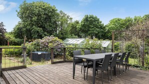 Een houten terras met een tafel en stoelen, omringd door groen en een kas.