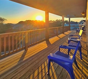 A wooden deck with blue chairs and a railing, overlooking a residential area during sunset. - OWNERS WEEKS OPEN⭐NEW HOT TUB⭐️Heated Pool, Linens, Wifi, Tennis Courts, Dogs  (Corolla)