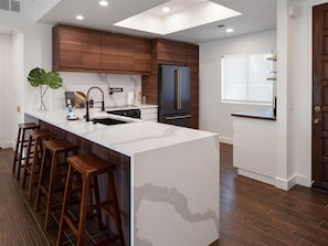 Una cocina moderna con una isla central, taburetes de madera, un refrigerador negro y una ventana con persianas.