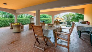 Eine Terrasse im Freien mit rotem Backsteinboden, überdacht von einer weißen Decke, mit Korbstühlen und einem Tisch.