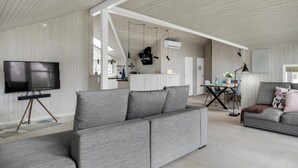 Un salon moderne avec un canapé d’angle gris, un téléviseur à écran plat posé sur un pied, un coin repas avec une table et des chaises en bois, et une cuisine en arrière-plan.