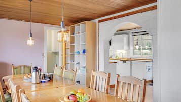 A dining area with a wooden table, chairs, and a fruit bowl. There is a kitchen visible through an archway.