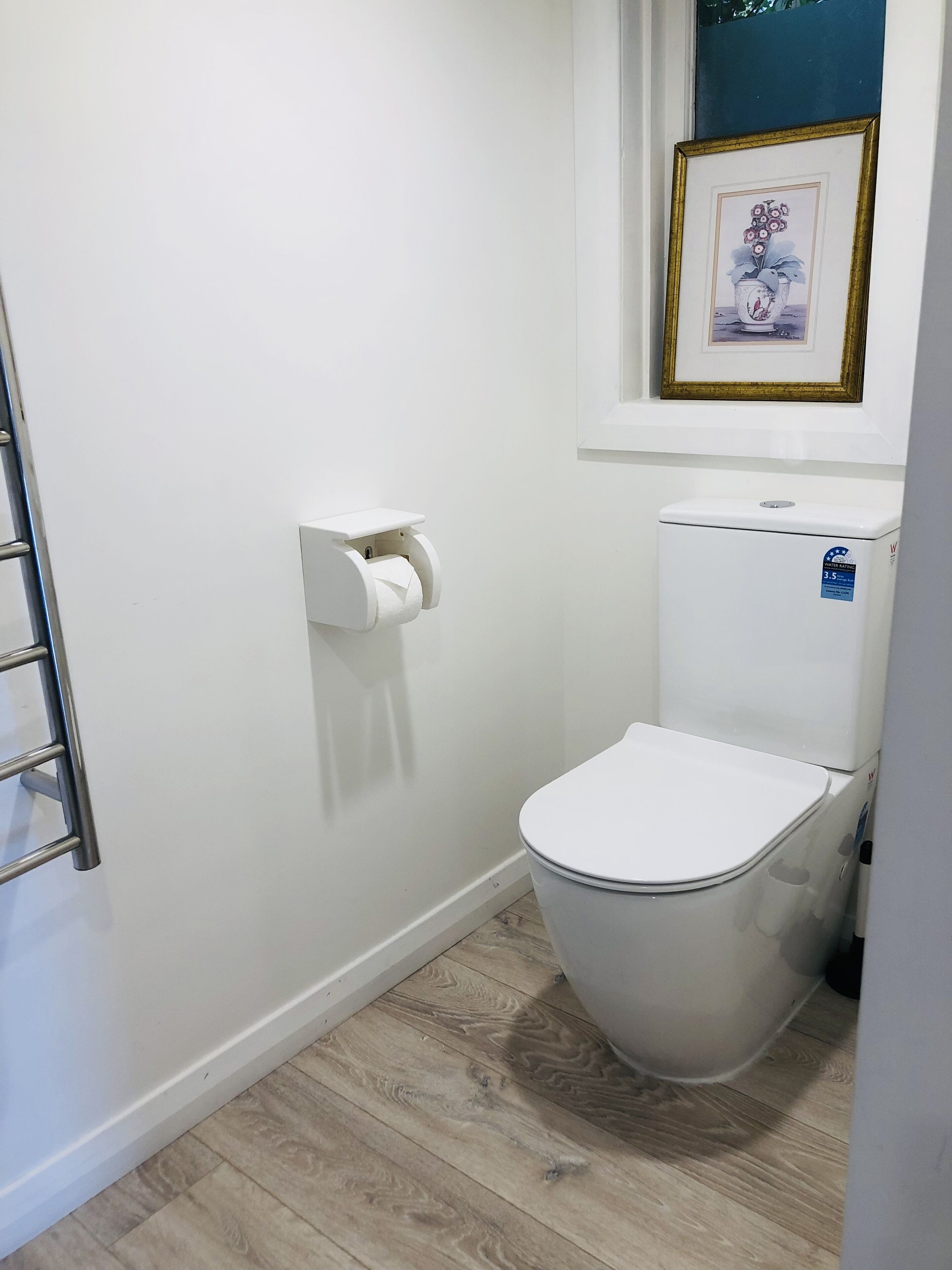 A small, clean bathroom with a toilet, a toilet paper holder, and a framed picture on the wall.