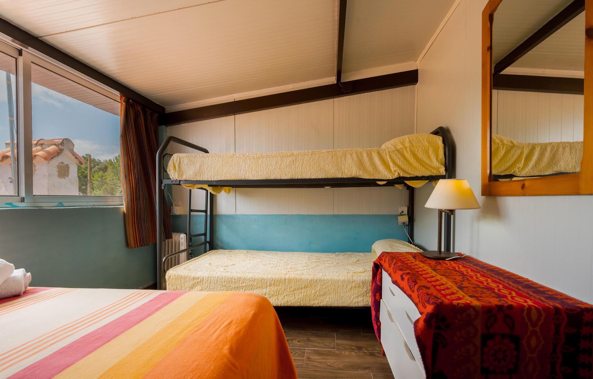 Ein Zimmer mit Etagenbetten, einem Nachttisch mit Lampe, einem Fenster mit Vorhängen und einem Bett mit gestreifter Decke.