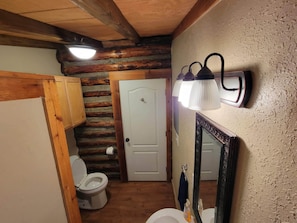 Une salle de bain avec un plafond en bois, des toilettes blanches, un lavabo avec un miroir et un luminaire fixé au mur.