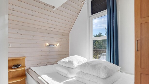 Un dormitorio con techo inclinado, una cama con ropa de cama blanca, un estante de madera y una ventana con cortinas.