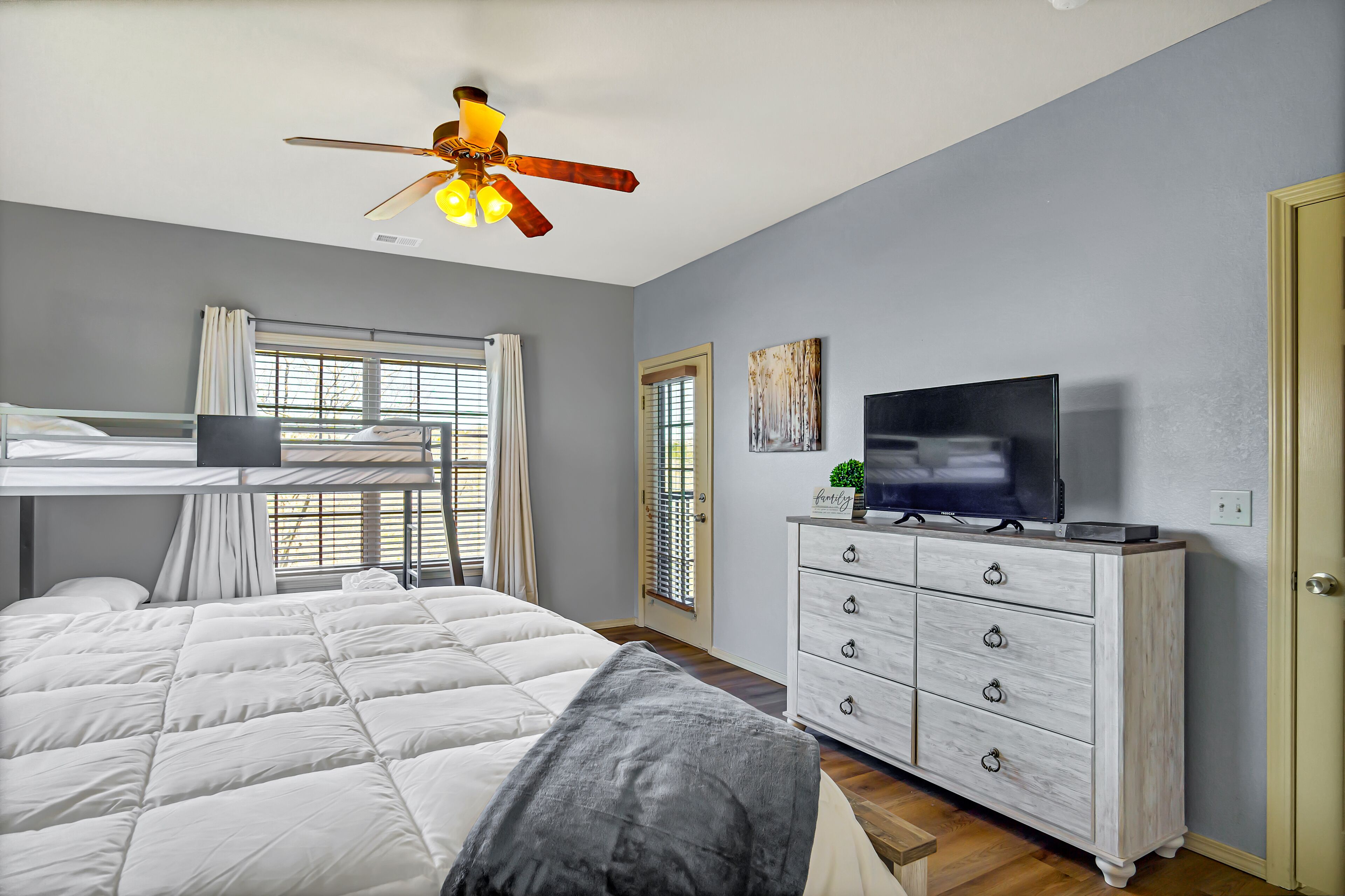 Une chambre avec un lit superposé, un ventilateur de plafond, une télévision, une commode et une fenêtre avec des rideaux.