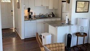 Une cuisine moderne avec des armoires blanches, des comptoirs en granit et un réfrigérateur encastré. On y trouve une chaise en osier avec un coussin et une petite table avec une lampe.