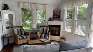 Un salon chaleureux, doté de meubles en osier, d’une cheminée et de grandes fenêtres offrant une vue sur l’extérieur.