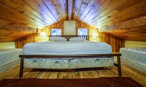 A cozy attic bedroom with a wooden ceiling, a large bed, two bedside tables with lamps, and a window on one wall.