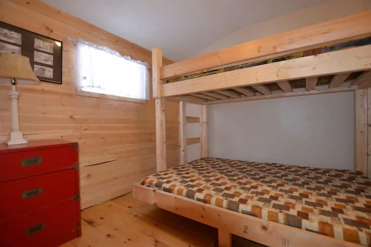 A small, wooden room with a bunk bed, a red dresser, and a window with curtains.