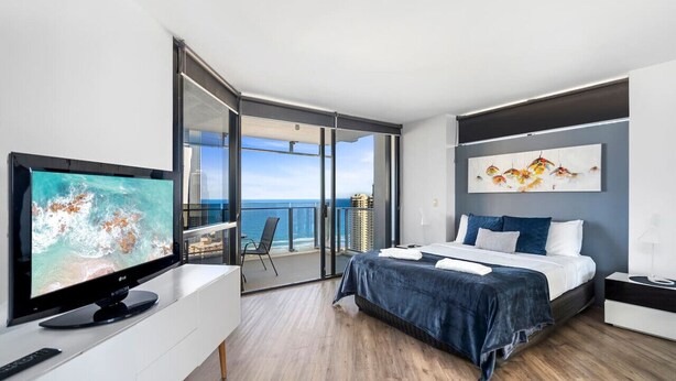 A modern hotel room with a large TV, a bed with blue bedding, a painting, and a balcony with a view of the sea. - Circle on Cavill Ocean View  Surfers Paradise (Surfers Paradise)