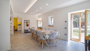 Een ruime eetruimte met een lange tafel gedekt voor een maaltijd, een keuken op de achtergrond en een schuifdeur naar een buitengebied.