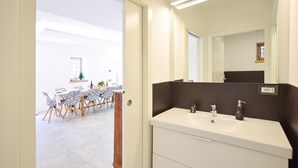 Een hotelkamer met een wastafel met dubbele wasbakken, een grote spiegel en een eethoek, zichtbaar door een geopende deur.