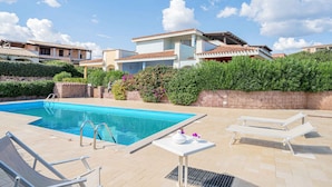 Una piscina con lettini e un tavolo, circondata da vegetazione e edifici.