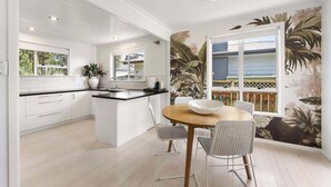 A modern kitchen with white cabinetry, a central island, and a dining area with a round table and chairs. - Five Palms Luxury Beach Escape (Mount Maunganui)
