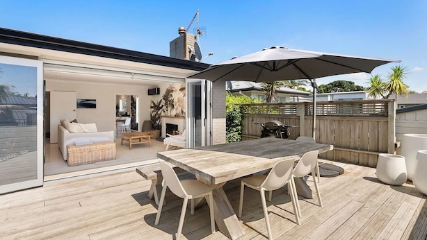 A wooden deck with a dining table and chairs, a sliding glass door leading to a living area with a sofa and a TV, and a wooden fence in the background. - Five Palms Luxury Beach Escape (Mount Maunganui)