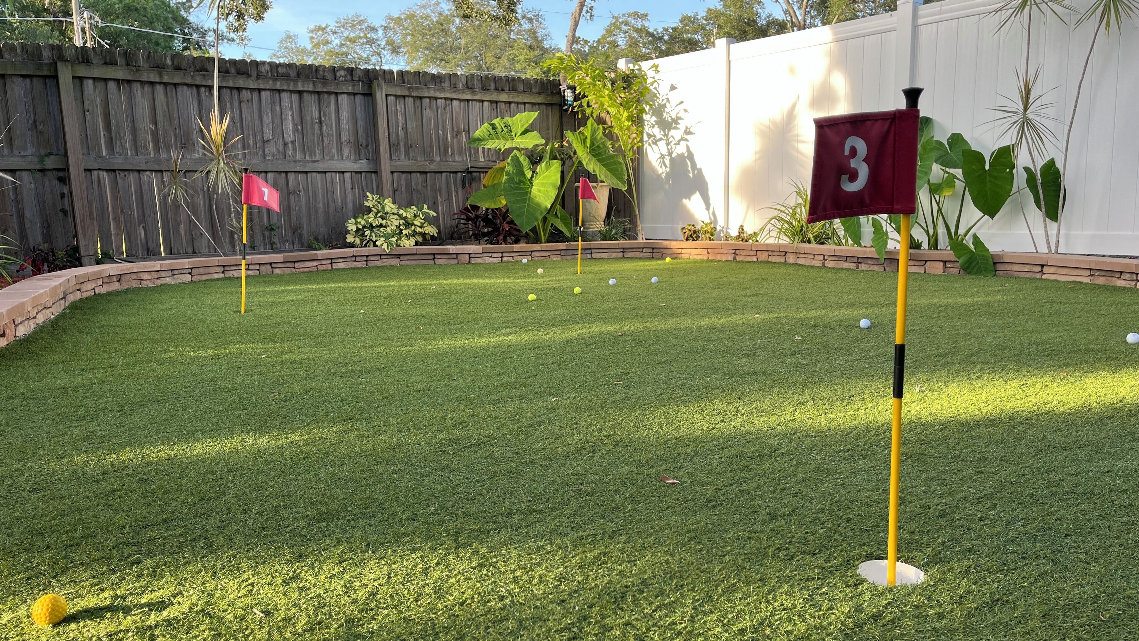 A backyard with a putting green, a wooden fence, and a white wall with a number 3 flag.
