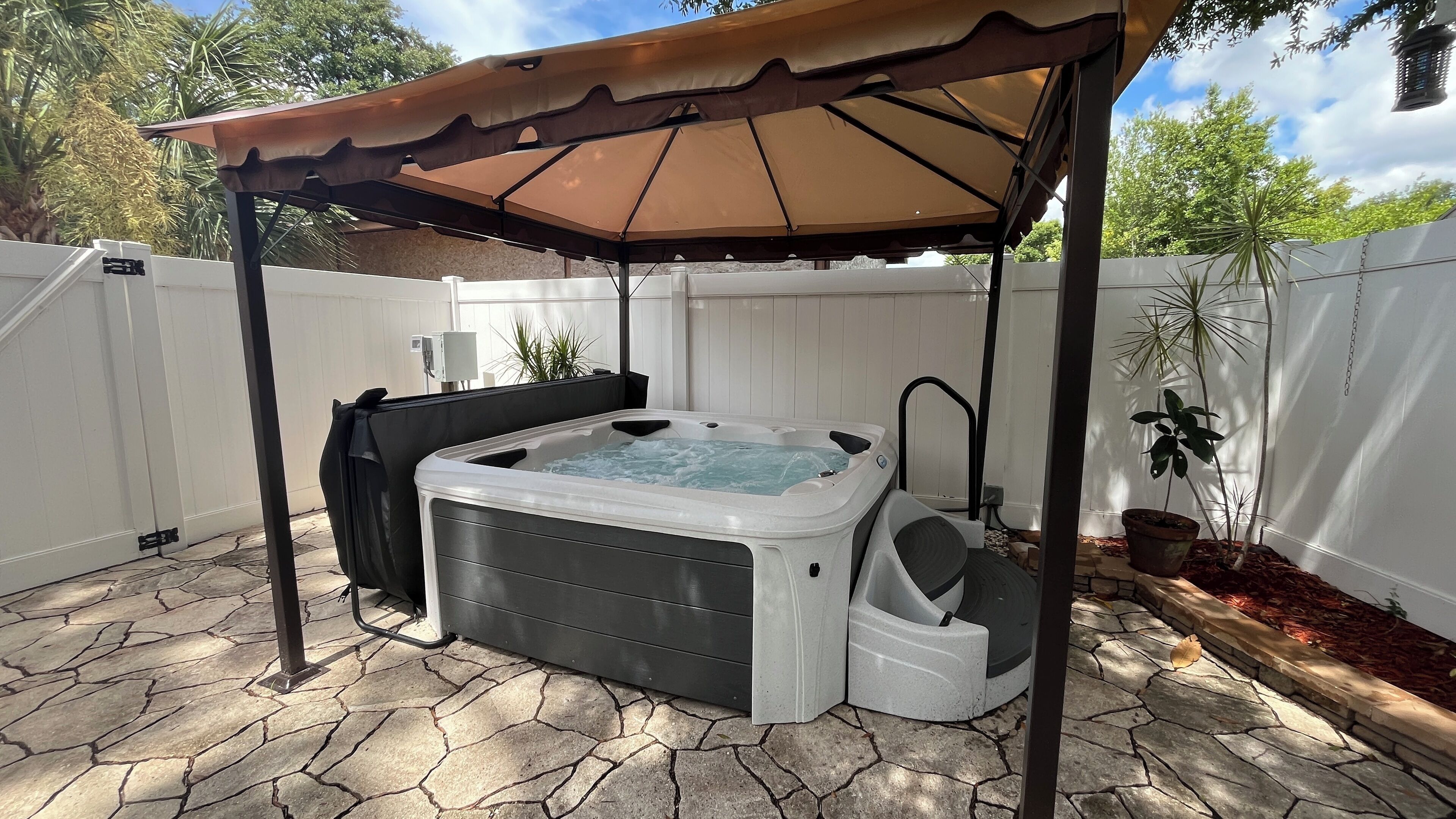 Un bain à remous installé sous une pergola, avec une terrasse en pierre, le tout entouré d’une clôture blanche et agrémenté de plantes en pot.