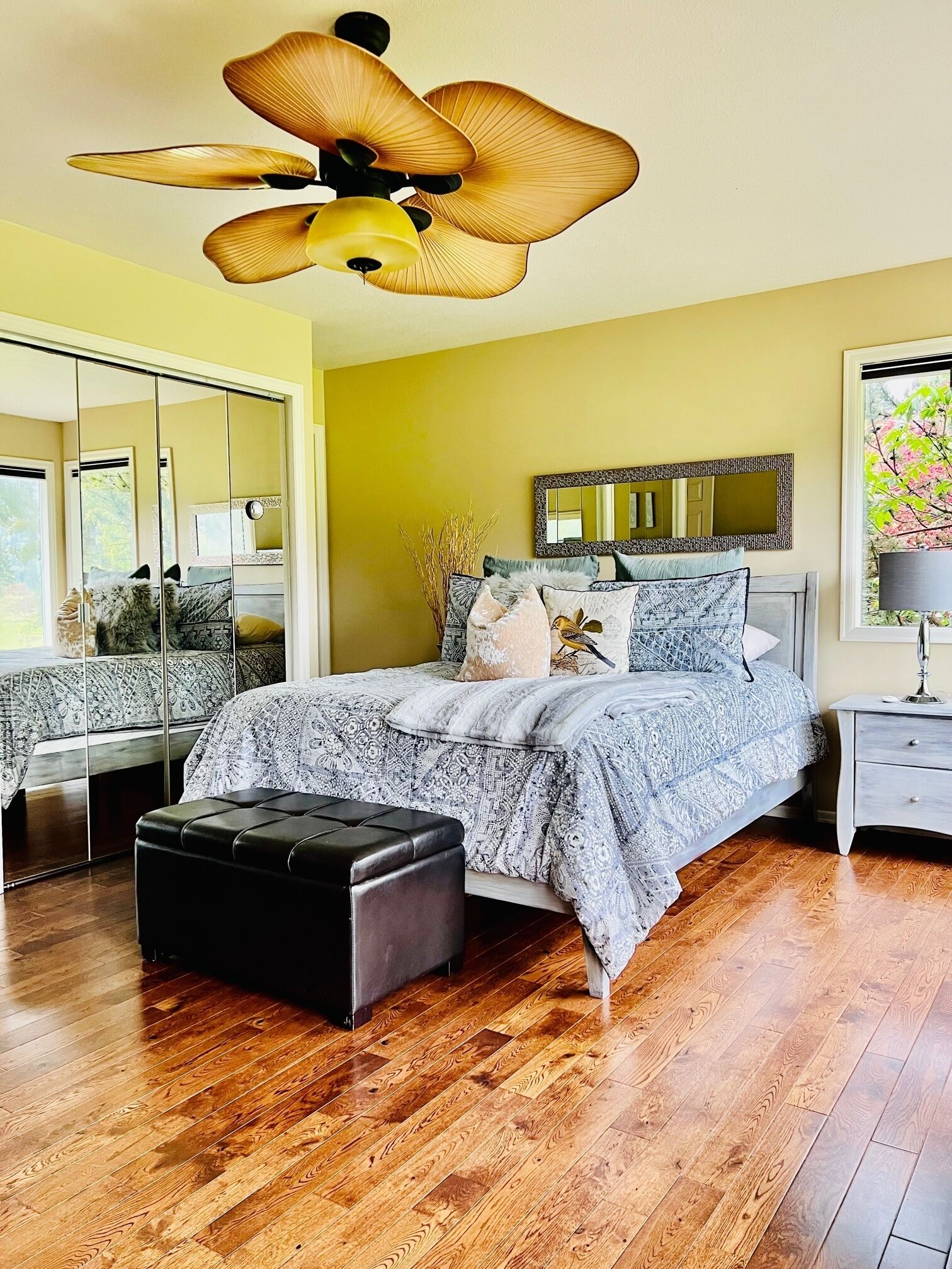 A bedroom with a bed, a ceiling fan, a nightstand, a mirror, and a window with a view of trees.
