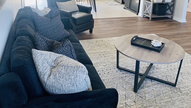 Un salon moderne avec un canapé noir, une table basse et un coin repas, la cuisine étant visible au fond.