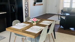 Un coin repas est aménagé avec une table en bois, dressée pour quatre personnes, entourée de chaises aux pieds en métal. Un réfrigérateur noir se trouve à gauche, et un téléviseur à écran plat est fixé au mur, au-dessus d’une étagère en bois.