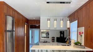 A modern kitchen with wooden paneling, stainless steel appliances, a central island with bar stools, pendant lighting, and a view of the adjacent living area. - New*Quiet Nature view #3; 1/1, pool, walk to beach (Fort Myers Beach)