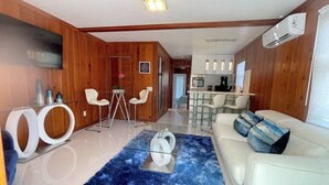A modern living room with a white sofa, a blue rug, and a glass coffee table. There is a kitchen area in the background with white cabinets and a microwave. - New*Quiet Nature view #3; 1/1, pool, walk to beach (Fort Myers Beach)