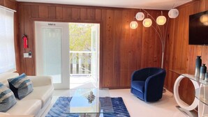 A modern living room with a glass coffee table, a blue armchair, and a television on a wooden wall. - New*Quiet Nature view #3; 1/1, pool, walk to beach (Fort Myers Beach)