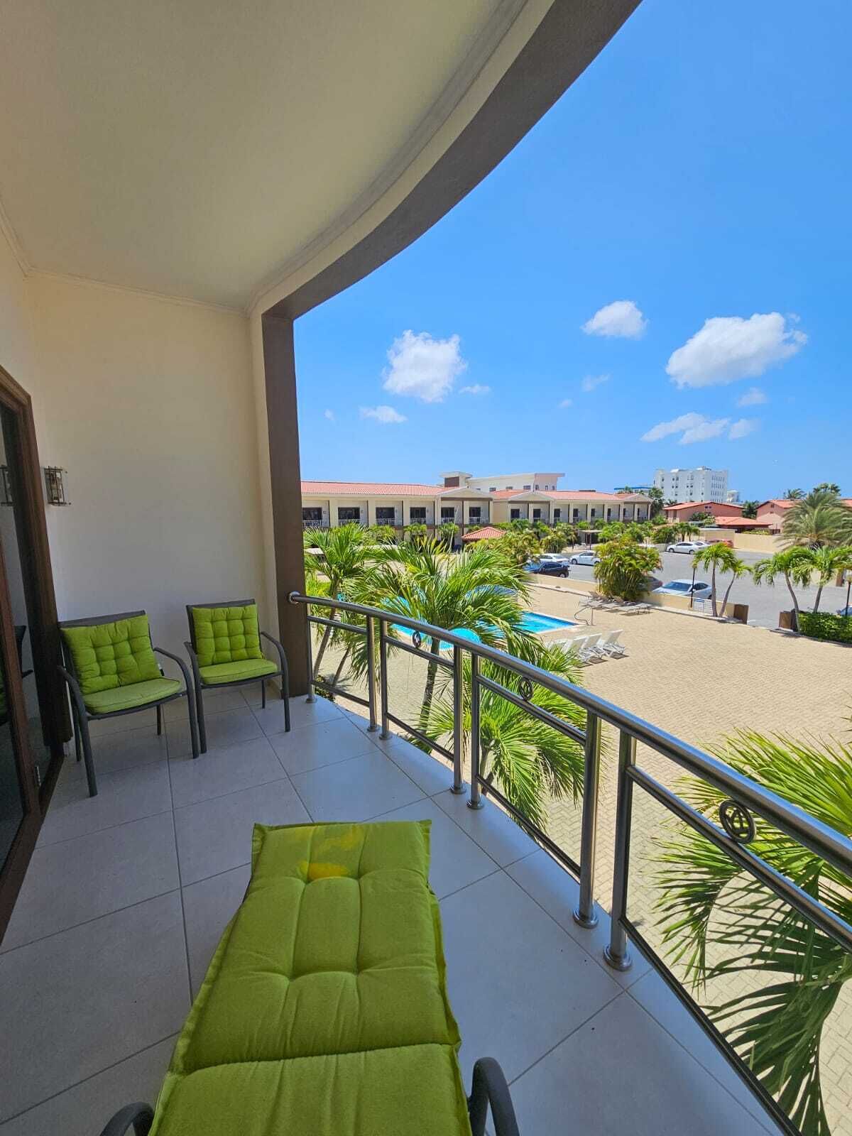 A balcony with green cushioned chairs, a pool, and a view of a building complex.
