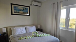 Un dormitorio con cama, mesita de noche, equipo de aire acondicionado, ventana con cortinas e imagen enmarcada en la pared.