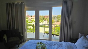 Habitación de hotel con cama, una silla y balcón con vista a palmeras y edificios.