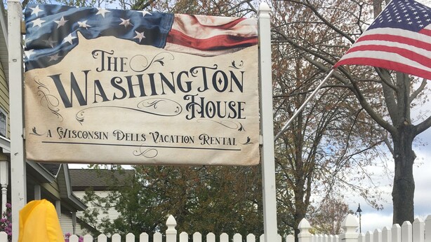 A sign for The Washington House, a Wisconsin Dells vacation rental, with a white picket fence and yellow daffodils in the foreground.