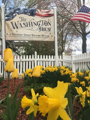 A sign for The Washington House, a Wisconsin Dells vacation rental, with a white picket fence and yellow daffodils in the foreground. - THE WASHINGTON HOUSE - A  4-Bedroom Home 1 Block from Downtown Wisconsin Dells (Wisconsin Dells)