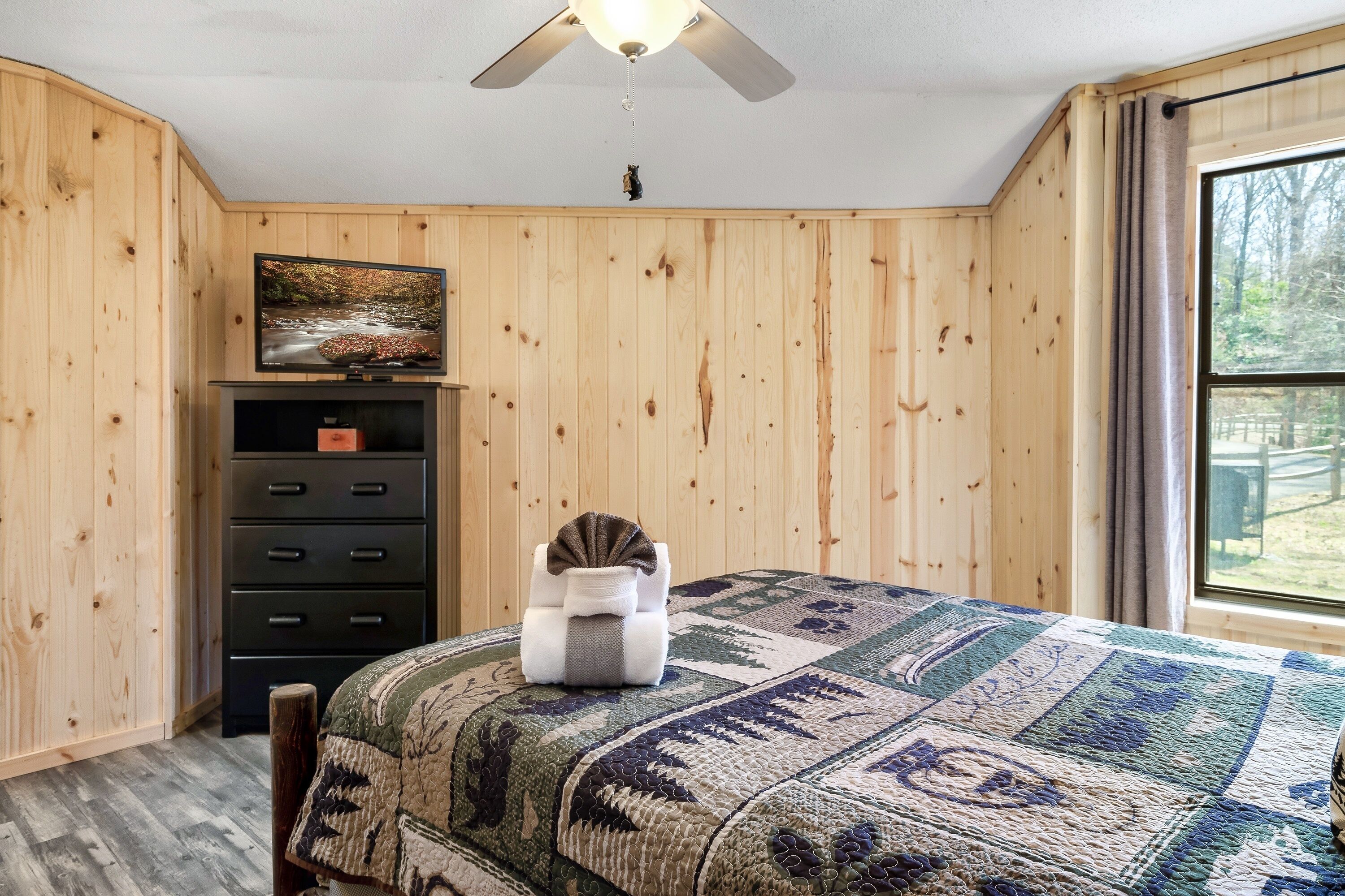 A bedroom with wooden walls, a bed with a patterned bedspread, a dresser, a television on the wall, and a window with a view of trees.
