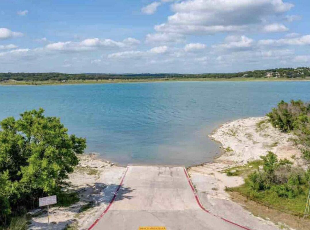 A beach with a concrete walkway leading to the water, surrounded by greenery and a clear sky.