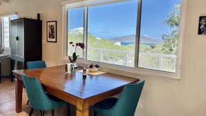 Ein Essbereich mit einem Holztisch, blaugrünen Stühlen, einer Topfpflanze und einem Fenster mit Blick auf das Meer und die Hügel.