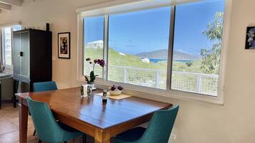 Un coin repas avec une table en bois, des chaises de couleur turquoise, une plante en pot et une fenêtre donnant sur la mer et les collines.