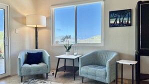 Ein Zimmer mit zwei gemusterten Sesseln, einem kleinen Tisch, einer Lampe und einem großen Fenster mit Meerblick.