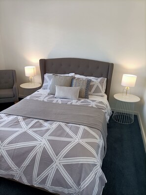 Un lit bien fait, recouvert d’une couvre-lit à motifs géométriques, deux tables de chevet avec des lampes et un fauteuil à revêtement gris.