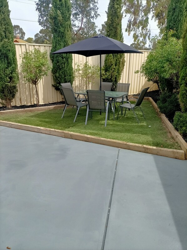 Un espace patio avec une table et des chaises sous un grand parasol sombre, entouré d’une pelouse et de haies bien entretenues.
