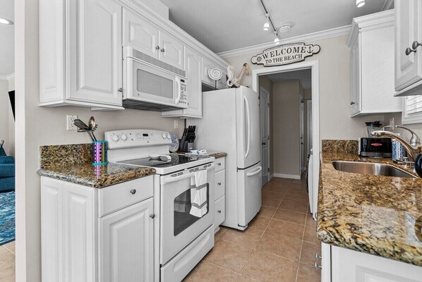 A kitchen with white cabinetry, a granite countertop, a microwave, an oven, a refrigerator, and a sign that reads 'WELCOME TO THE BEACH'. - Newport Beach 1 bedroom, 1 bath + Sleeper Sofa Bed - Home/Beach/Garage/Patio (Newport Beach)