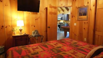 A wooden room with a bed, a television, a lamp, and a door leading to a kitchen.