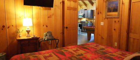 Una habitación de madera con una cama, un televisor, una lámpara y una puerta que da a una cocina.