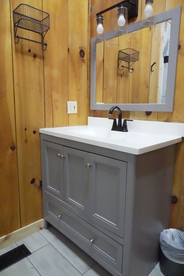 A bathroom with a wooden wall, a white sink, a mirror with lights, and a trash bin. - Moonshine Holler (Sevierville)