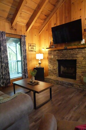 A cozy living room with wooden walls, a stone fireplace, a flat-screen TV mounted above it, and a comfortable seating area with a sofa and a coffee table. - Moonshine Holler (Sevierville)