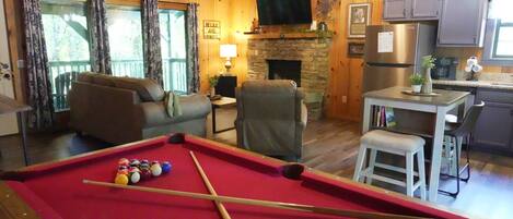 A cozy room with a pool table, a fireplace, a television, and a kitchen area.